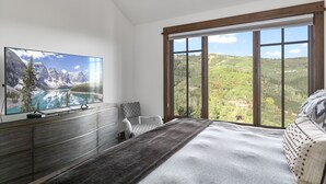 A bedroom with a large window, a bed, a TV on a wooden cabinet, and a view of mountains and a lake.