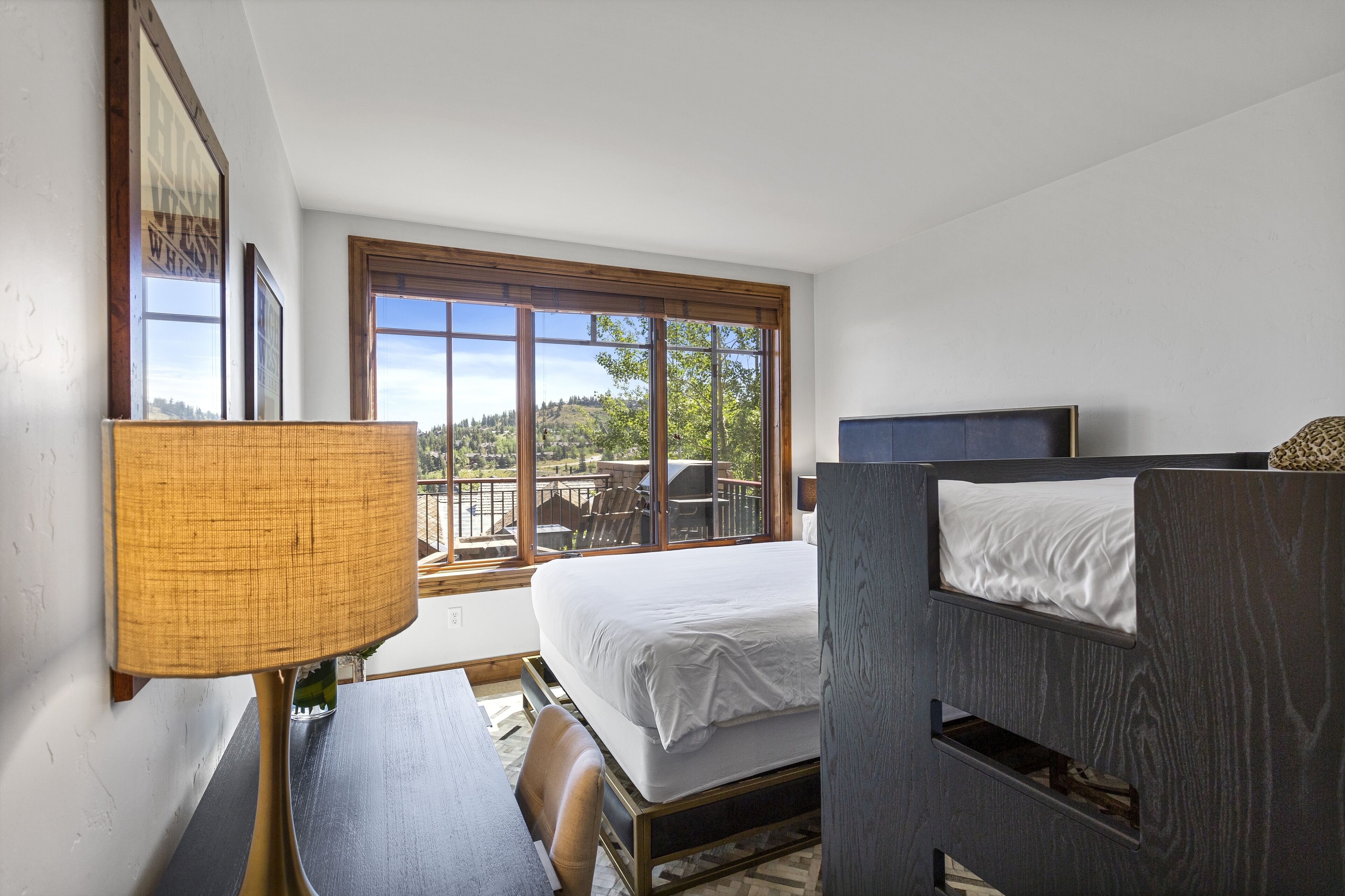 A bedroom with a bed, a desk, a chair, a window with a view of the outdoors, and a lamp.