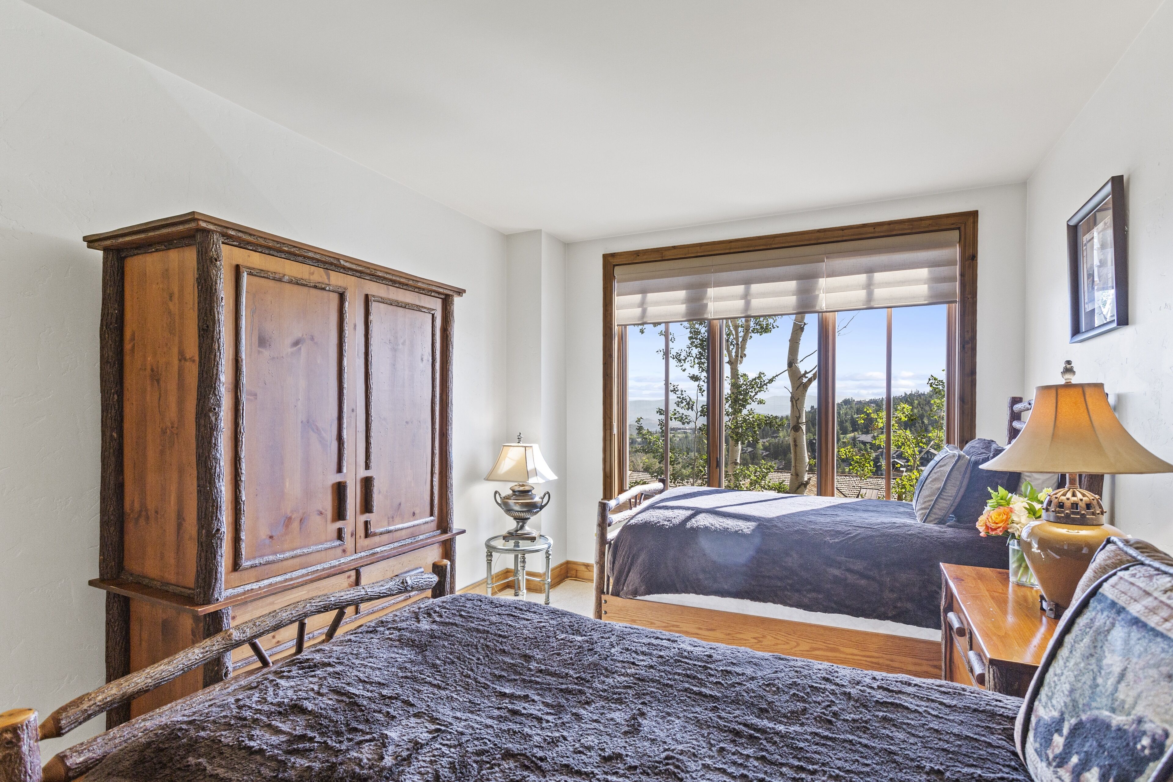 Une chambre avec deux lits, une armoire en bois, une table de chevet avec une lampe, et une vue sur l’extérieur grâce à de grandes fenêtres.