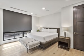 Ein modernes Schlafzimmer mit einem großen Bett, Nachttischen, einer Bank und Blick auf die Straße durch ein Fenster.