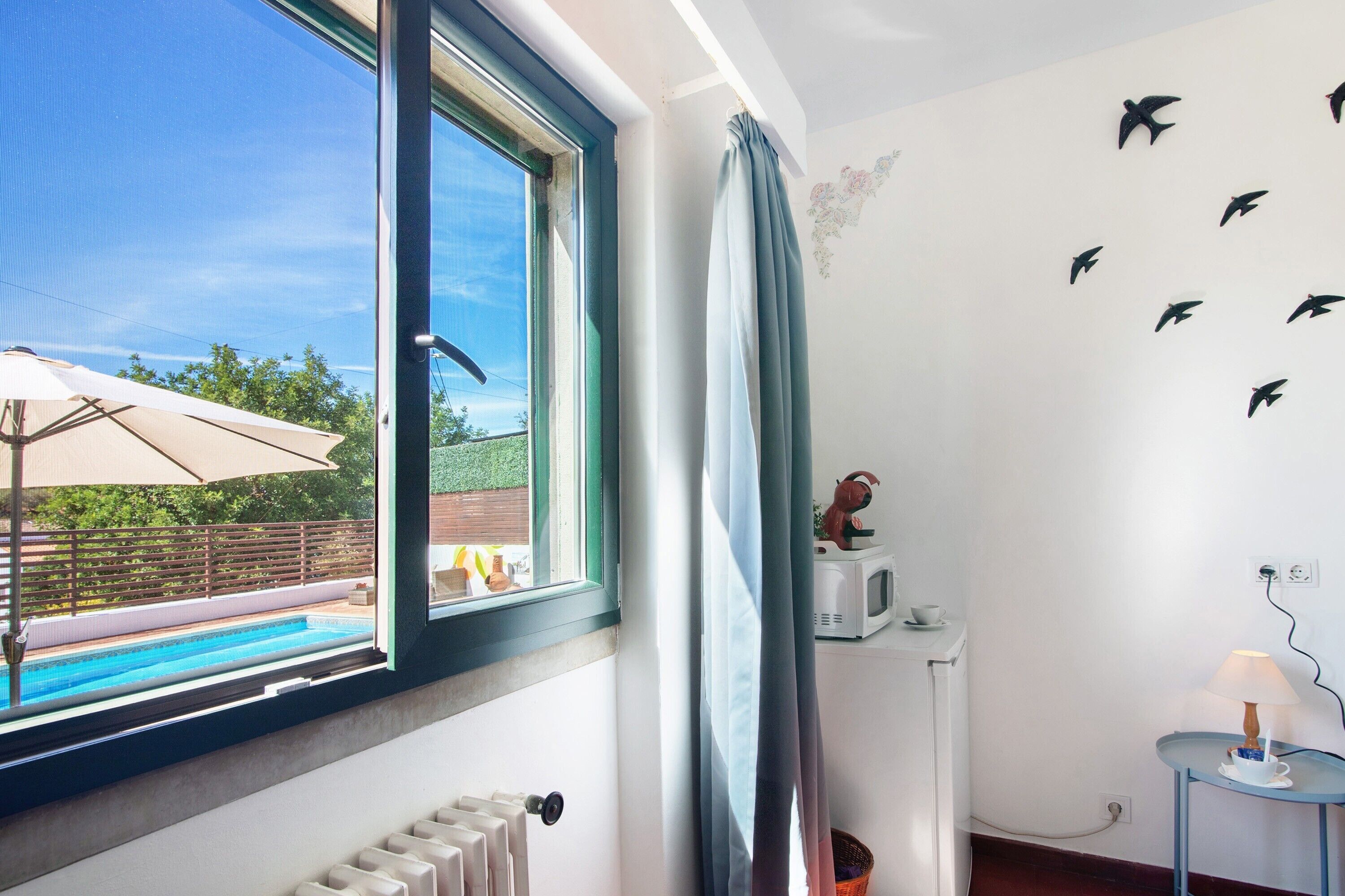 Une chambre avec une fenêtre donnant sur une piscine, un radiateur, une petite table avec une lampe et un mur décoré d’autocollants représentant des oiseaux.