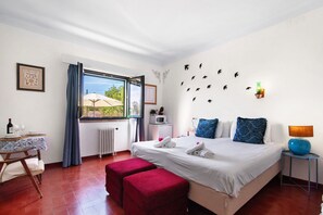 Ein Schlafzimmer mit einem Bett, einem Tisch mit Flasche und Gläsern, einer roten Bank, einem Fenster mit Vorhängen, einem Heizkörper, einer Wandleuchte und einer Wanddekoration mit Vogelmotiven.