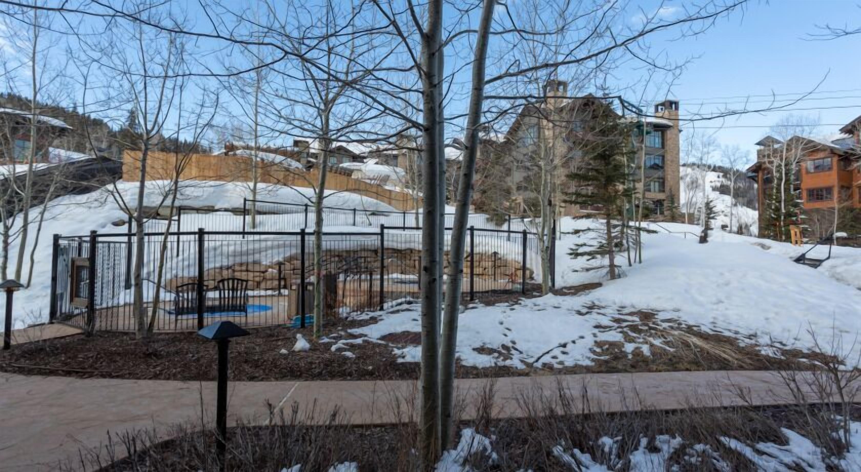 A snow-covered outdoor area with a fenced playground, trees, and residential buildings in the background.