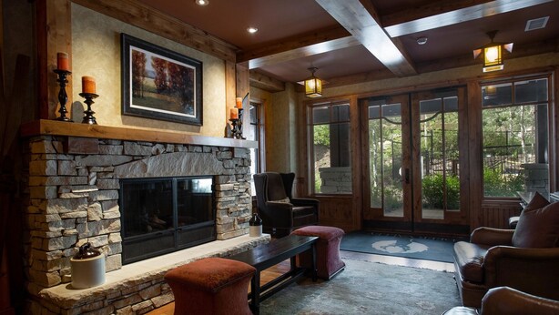A stone fireplace with a television, a bench, and two chairs in a room with wooden beams and large windows.