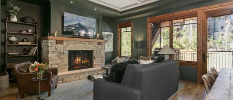 Una sala de estar acogedora con chimenea de piedra, un televisor de pantalla plana grande, un sofá gris y una mesa de comedor de madera.