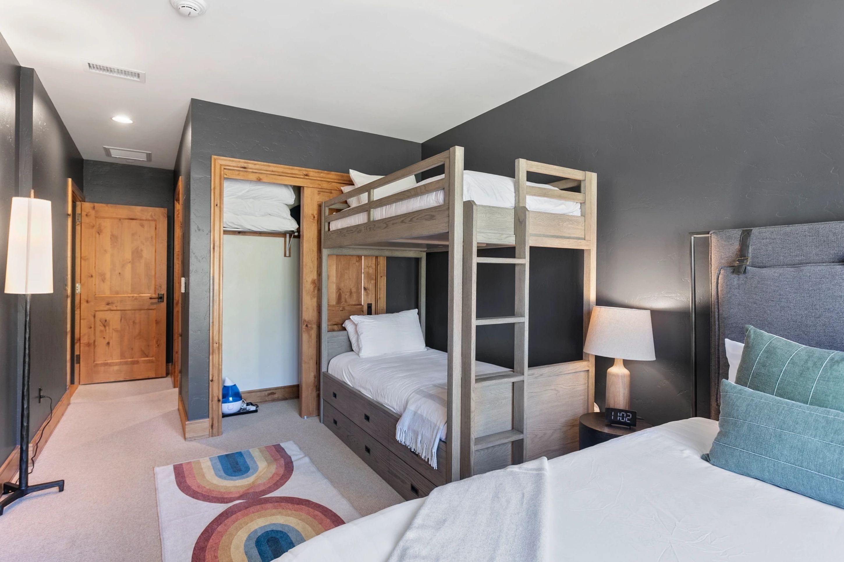 Quarto com beliches, porta de madeira, mesa de cabeceira com abajur e tapete colorido.