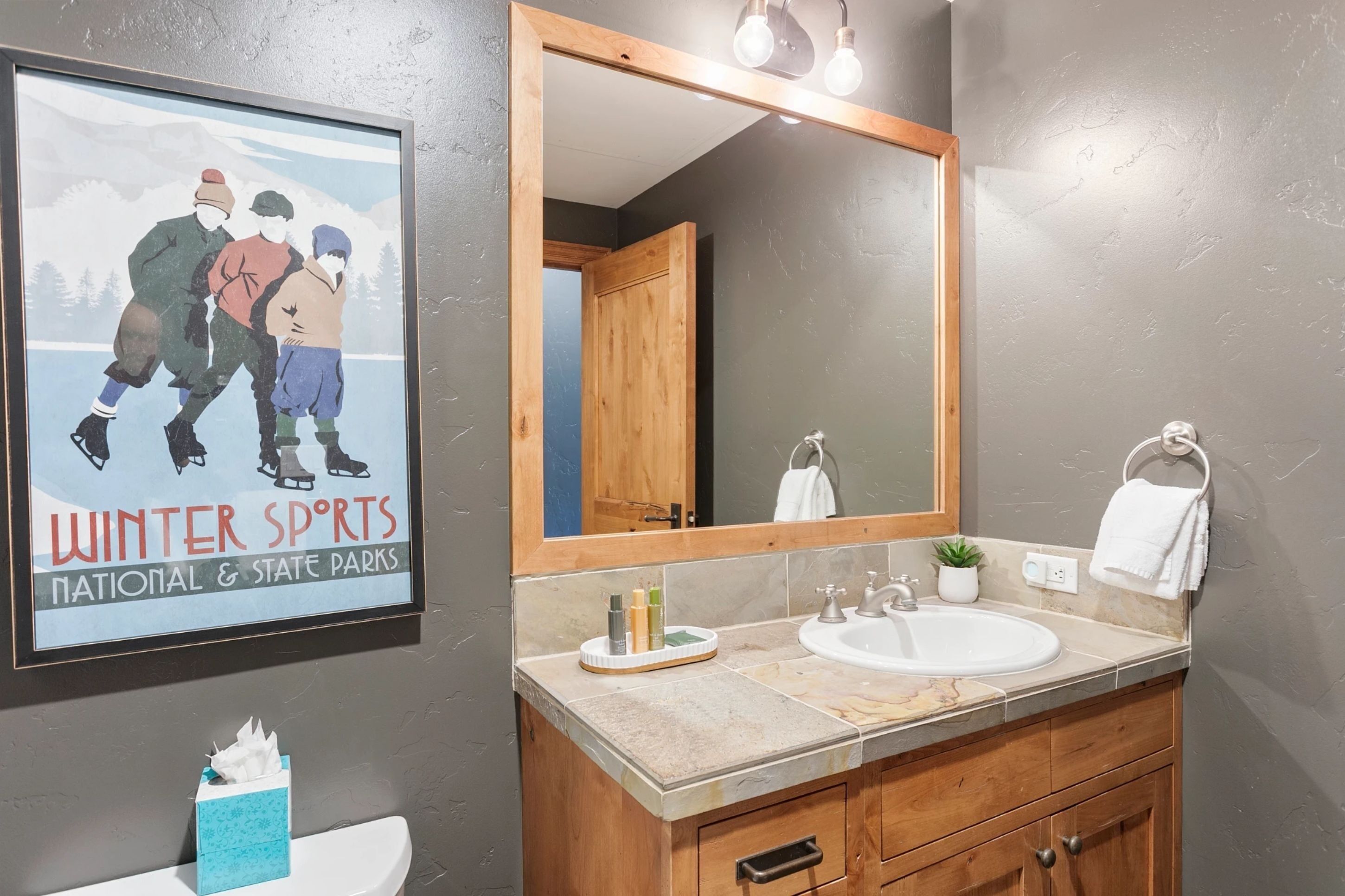 Un baño con un espejo grande, un lavabo, un perchero para toallas y un cartel con atletas de deportes de invierno.