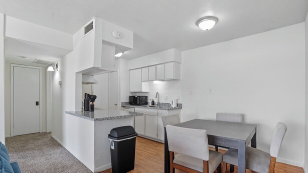 A compact kitchen with white cabinets, a black trash bin, and a dining area with a gray table and white chairs. - Apt. In college station TX. Mins to major hospitals and A&M U (College Station)