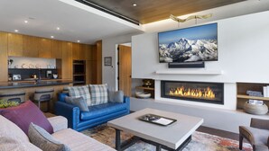 A modern living room with a fireplace, a mounted TV, and a kitchen area in the background. - One Empire Pass at Deer Valley Resort - Three Bedroom Residence #402 3 Condo (Park City)