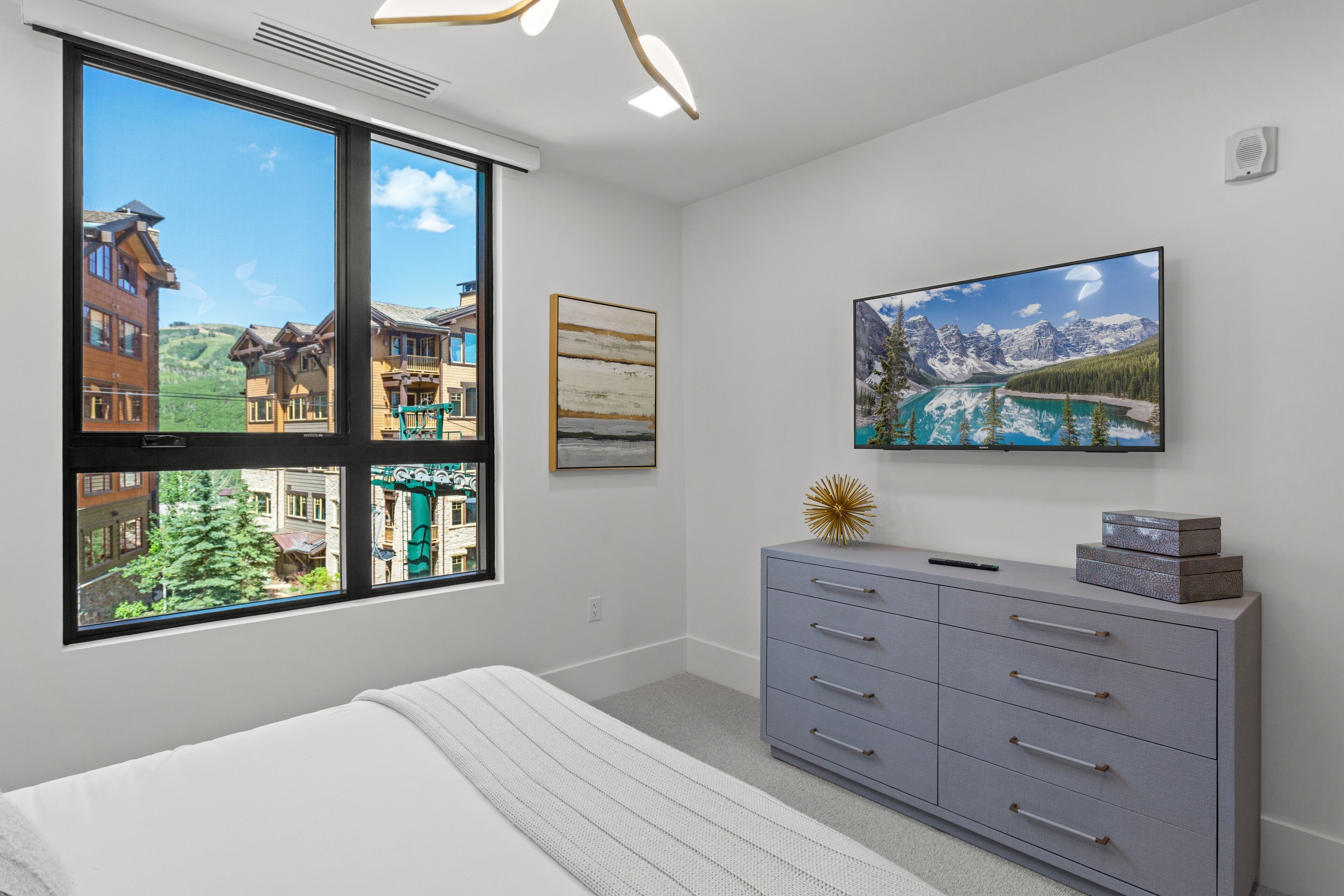 A bedroom with a large window offering a view of buildings and trees, a bed, a dresser, and two framed artworks.