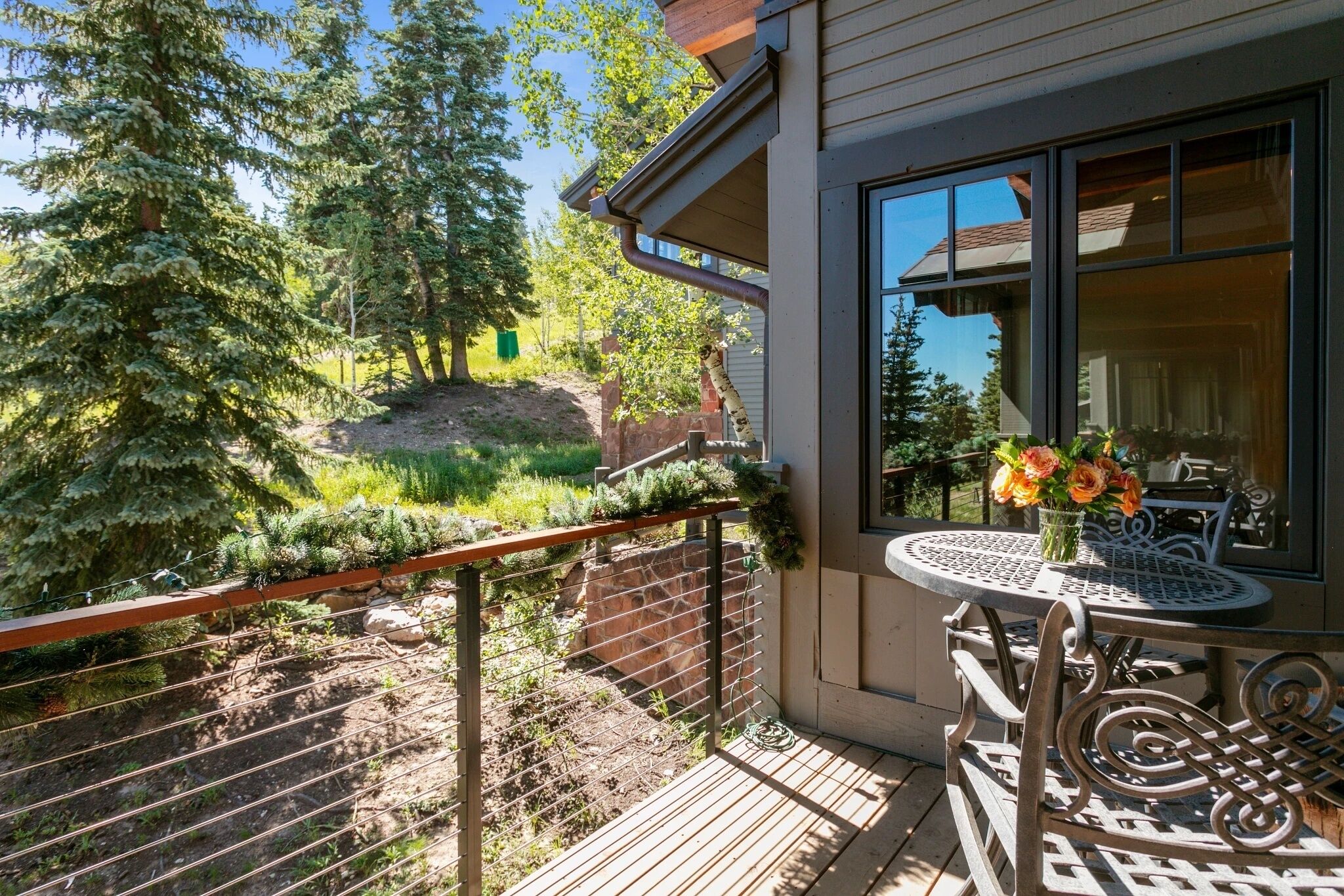 A balcony with a view of a forested area, a table with a vase of flowers, and a window with a view of a building.