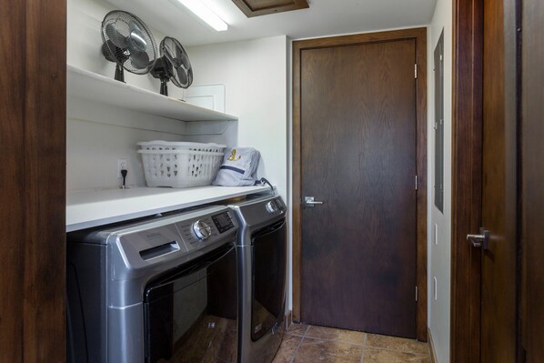 A laundry room with two washing machines, a dryer, a shelf with laundry baskets, and two wall fans.