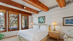 A bedroom with a large bed, wooden beams, a window with a view of trees, a bedside table with a lamp, and a framed picture on the wall.