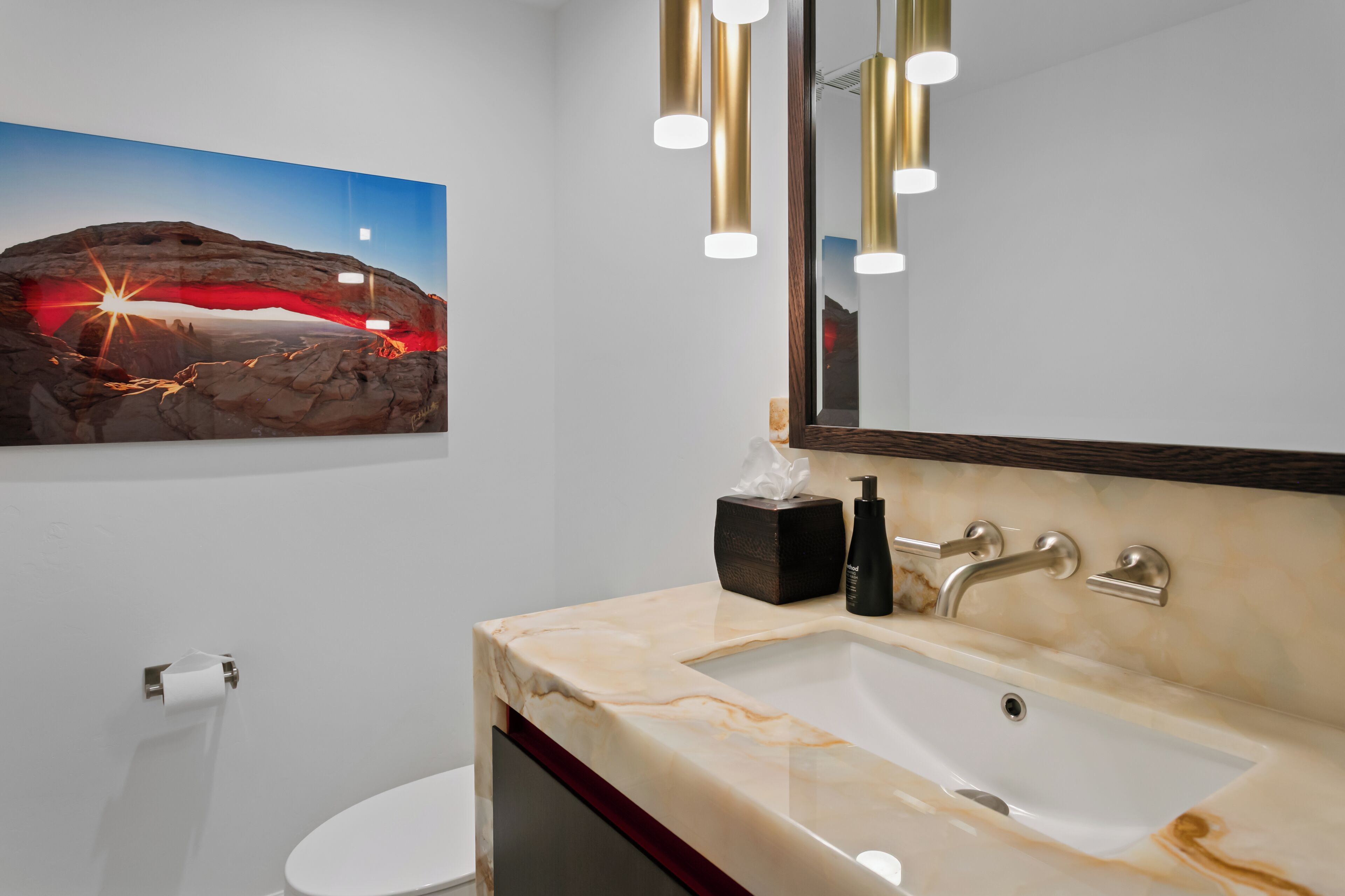 A bathroom with a marble sink, a mirror, a towel dispenser, and a painting of a landscape.