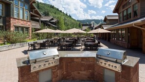 An outdoor patio with stone barbecue grills, tables, and umbrellas, surrounded by lodge buildings and a mountainous backdrop. - The Lodges at Deer Valley Hotel Room (Park City)