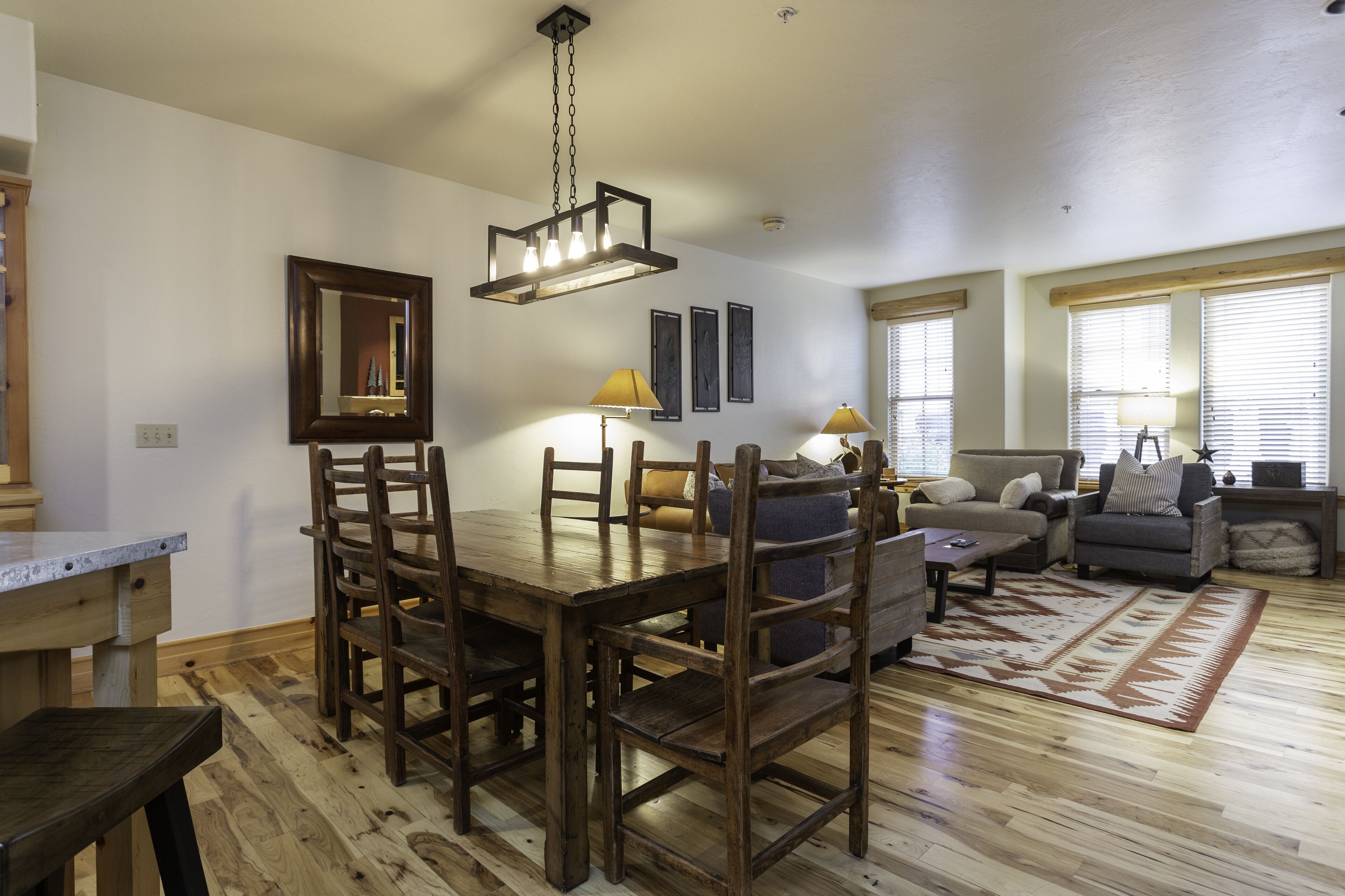 A dining area with a wooden table and chairs, a hanging light fixture, and a sofa in the background.