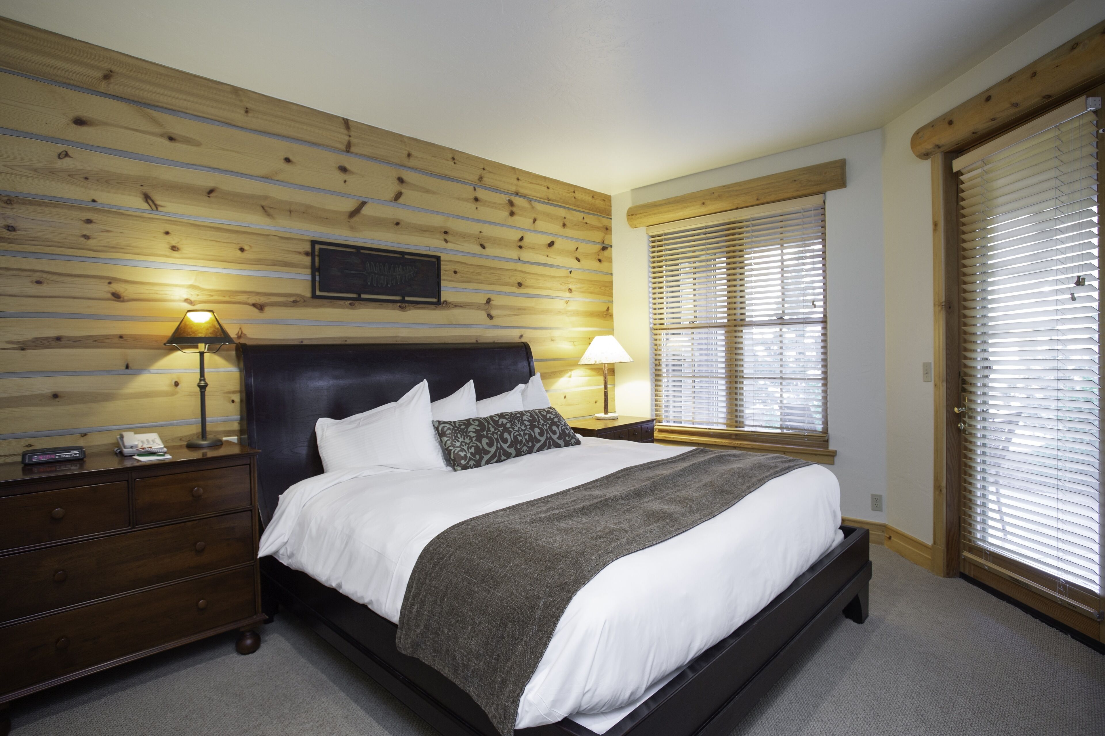 Un dormitorio con pared de listones de madera, una cama con ropa de cama blanca, dos lámparas de noche, una mesita de noche y una ventana con persianas.