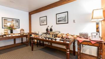 Une pièce avec une longue table en bois, dressée pour un buffet, où sont disposés divers plats et ustensiles. Les murs sont ornés de tableaux encadrés, une lampe se trouve sur une table d’appoint et un luminaire est suspendu au plafond.