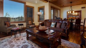 A spacious living room with a large window, a leather sofa, a wooden coffee table, and a dining area with a chandelier.