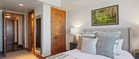 Un dormitorio con una cama grande, una puerta de madera, una mesita de noche con lámpara y un cuadro en la pared.
