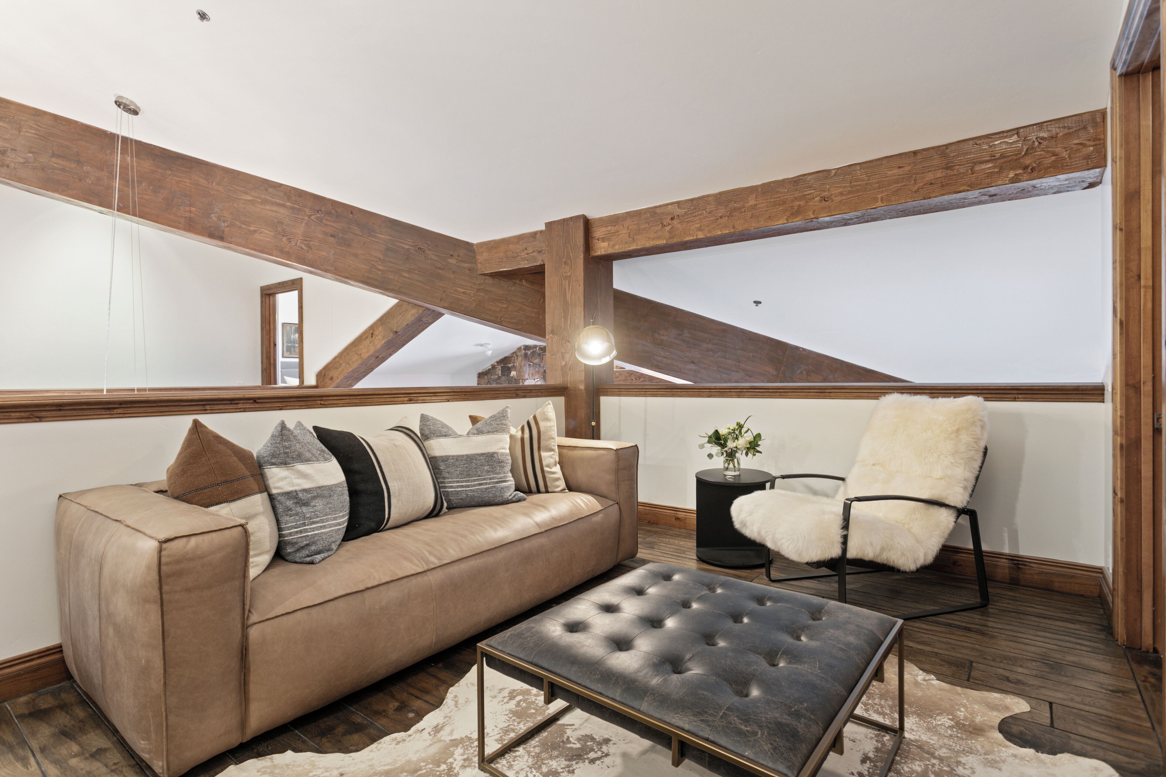 A modern loft bedroom with exposed wooden beams, a leather sofa, a tufted ottoman, a fluffy chair, and a small side table.