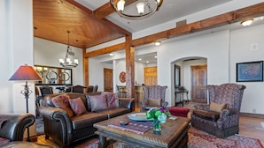 A spacious living room with a large sofa, two armchairs, and a coffee table. The room features exposed wooden beams, a chandelier, and a large mirror. - Grand Lodge - 3 Bedroom Condo Deluxe #206 - Ski-in/out by Deer Valley Resort (Park City)