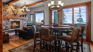 A cozy living room with a stone fireplace, wooden beams, a large dining table with chairs, and a view of trees through large windows. - Grand Lodge - 3 Bedroom Condo Deluxe #206 - Ski-in/out by Deer Valley Resort (Park City)