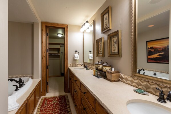 Un baño con lavamanos doble, tocador y un espejo grande. Hay una puerta de madera que conduce a otra habitación, y una alfombra en el suelo.