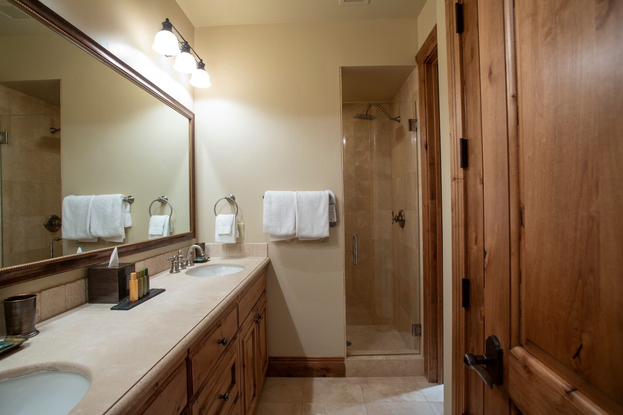 A bathroom with a large mirror, two sinks, a shower, and wooden cabinets.