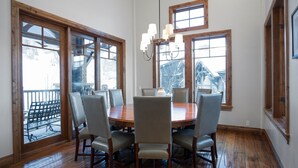 A dining room with a wooden table and six chairs, large windows, and a chandelier. - Grand Lodge - Five Bedroom Condo #402 - Ski-in/ski-out by Deer Valley Resort (Park City)