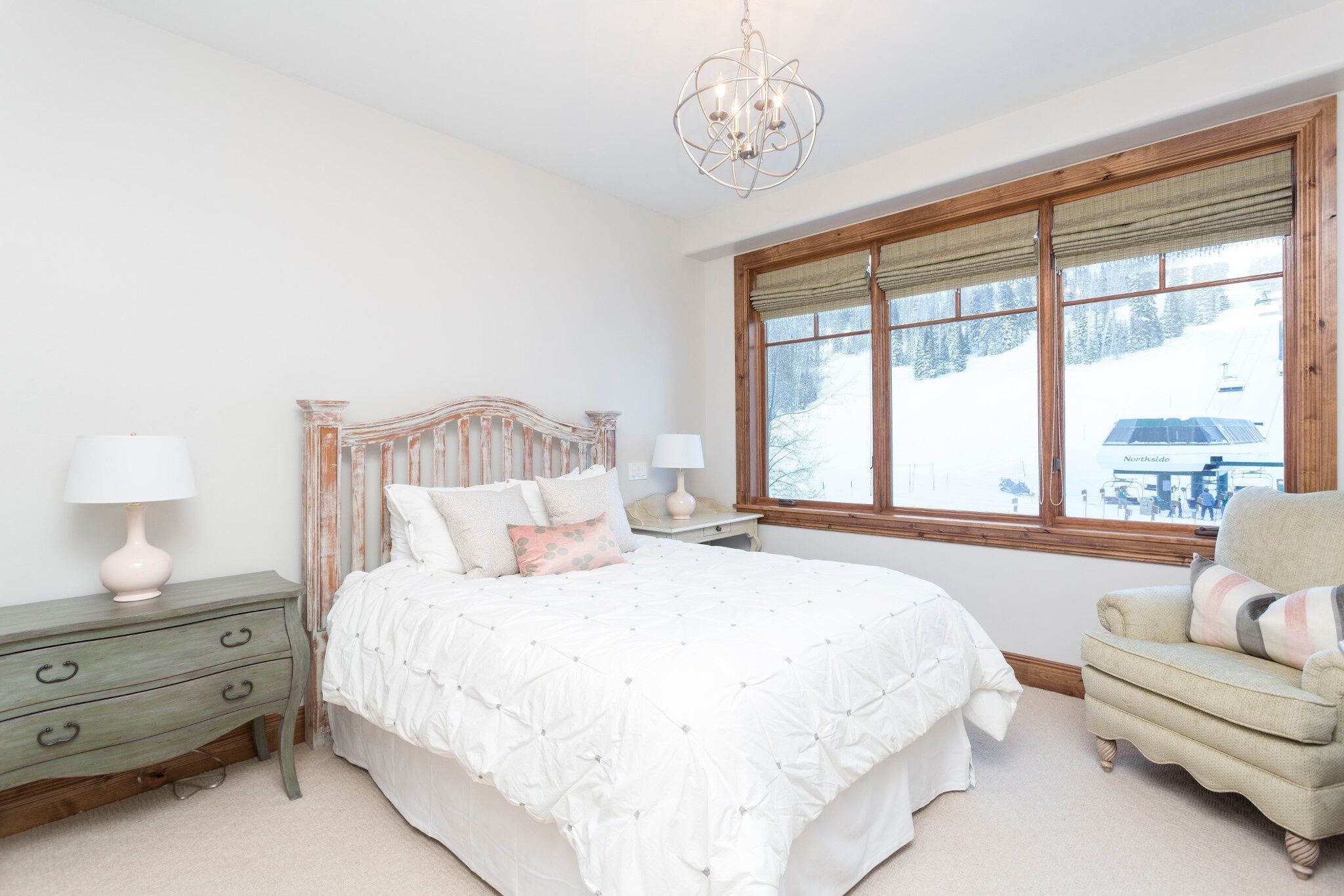 A bedroom with a large bed, a wooden headboard, a window with a view of a snowy landscape, a chair, and a nightstand with a lamp.
