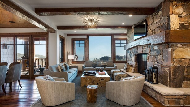 A spacious living room with a stone fireplace, large windows, and a wooden ceiling.