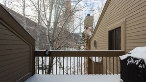 A snow-covered balcony with a metal railing, a covered car, and a house with beige siding and a chimney.