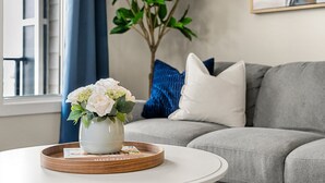 A living room with a grey sofa, a white coffee table with a tray, a vase with flowers, and a plant in the background. - Lion s Den - 3bed - 5guest - 2 Smarttv - Hospital (Calgary)