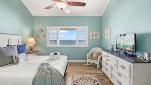 A bedroom with a bed, a ceiling fan, a television, a dresser, and a window with shutters.