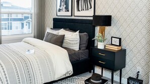 Een slaapkamer met een bed, nachtkastje, lamp, ingelijste foto’s en een raam met uitzicht op gebouwen.
