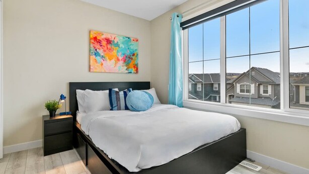 A bedroom with a large window, a bed with white bedding, a nightstand with a lamp, and a colorful abstract painting on the wall. - Golden Retreat - Games Movie Room - 8BD - 5bath (Calgary)