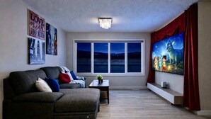 A living room with a sectional sofa, a small table with a plant, a large window with a view of the night sky, and a wall with framed pictures and a quote. - Golden Retreat - Games Movie Room - 8BD - 5bath (Calgary)