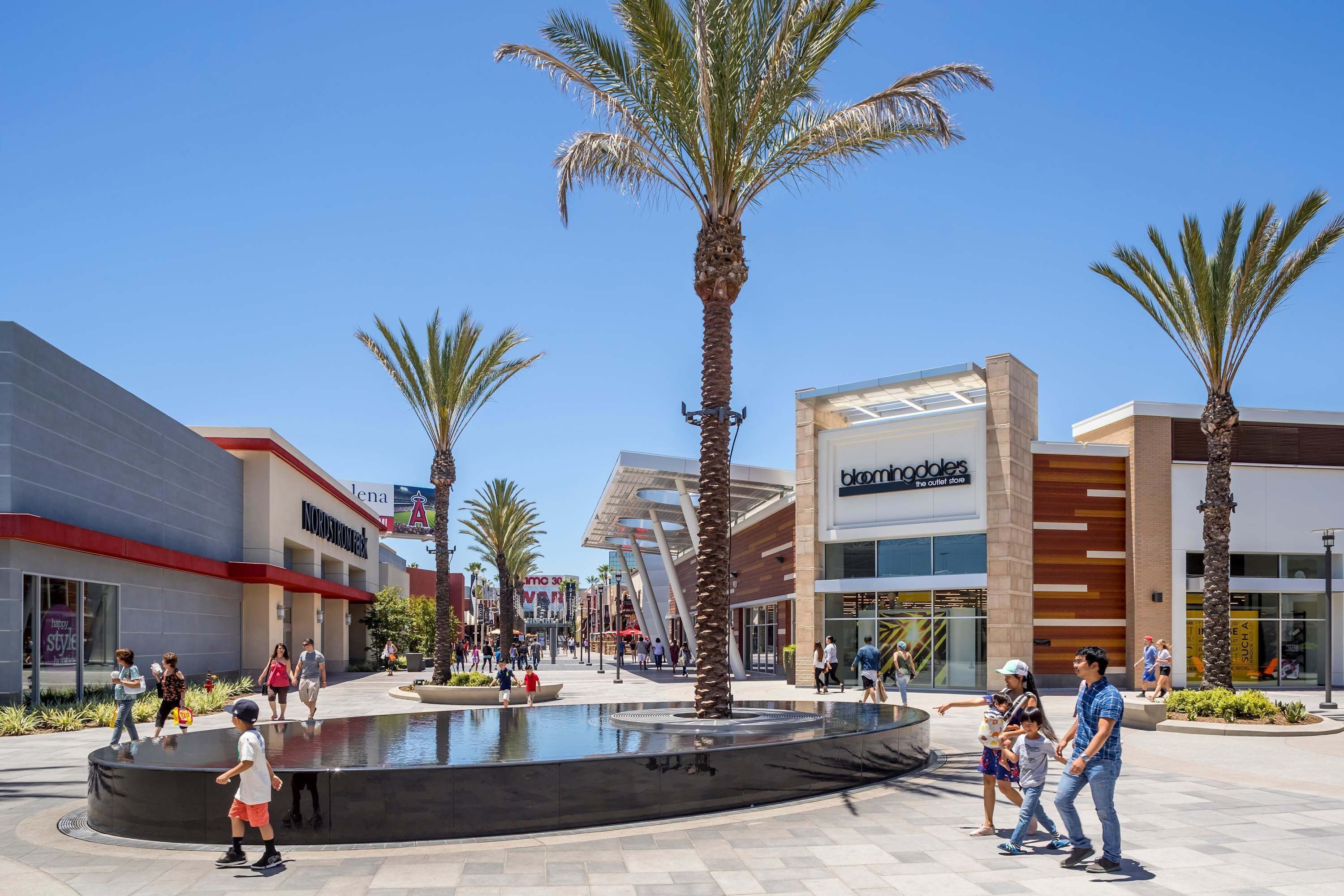A shopping plaza with palm trees, a fountain, and modern buildings.