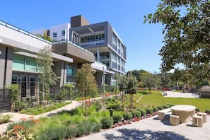 A modern building with a flat roof, large windows, and a landscaped outdoor area with a seating area and trees. - Pacific Paradise 10 Min From Disneyland With Pool GYM (Santa Ana)
