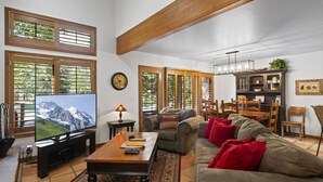 A spacious living room with a large flat-screen TV, wooden beams, a dining area, and a kitchen with wooden cabinets.