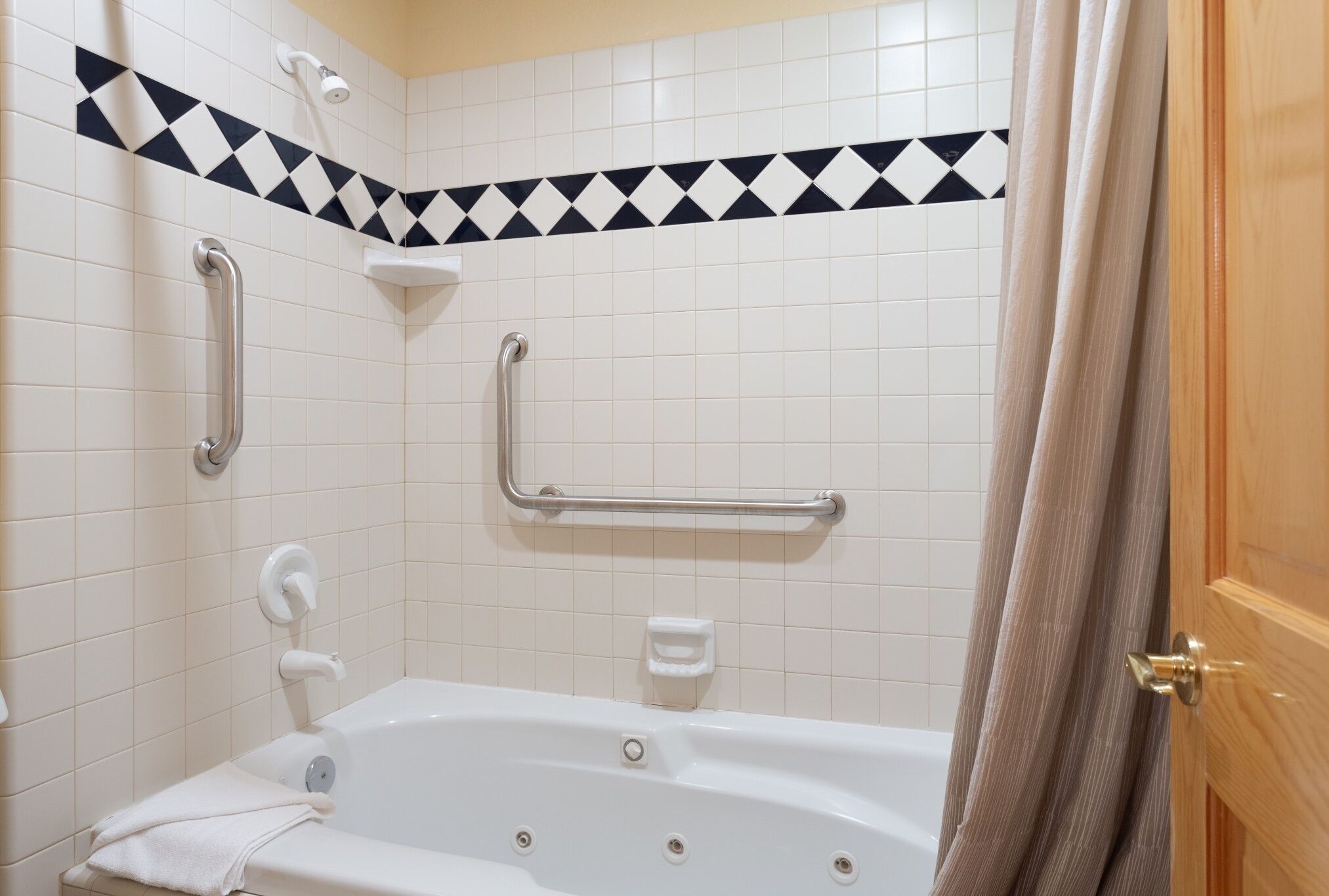 A bathroom with a white bathtub, grab bars, a showerhead, and a towel on the floor.