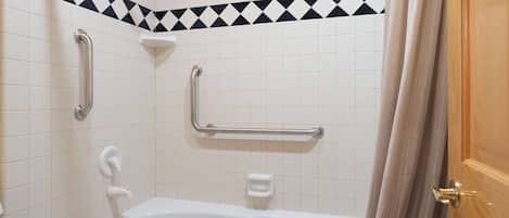 A bathroom with a white bathtub, grab bars, a showerhead, and a towel on the floor.