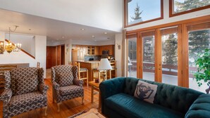 A spacious living area with a green sofa, patterned armchairs, a chandelier, and a view of a wooden deck and trees through large windows. - Ridge at Deer Valley Resort - Three Bedroom Condo with Hot Tub #16 3 Condo (Park City)