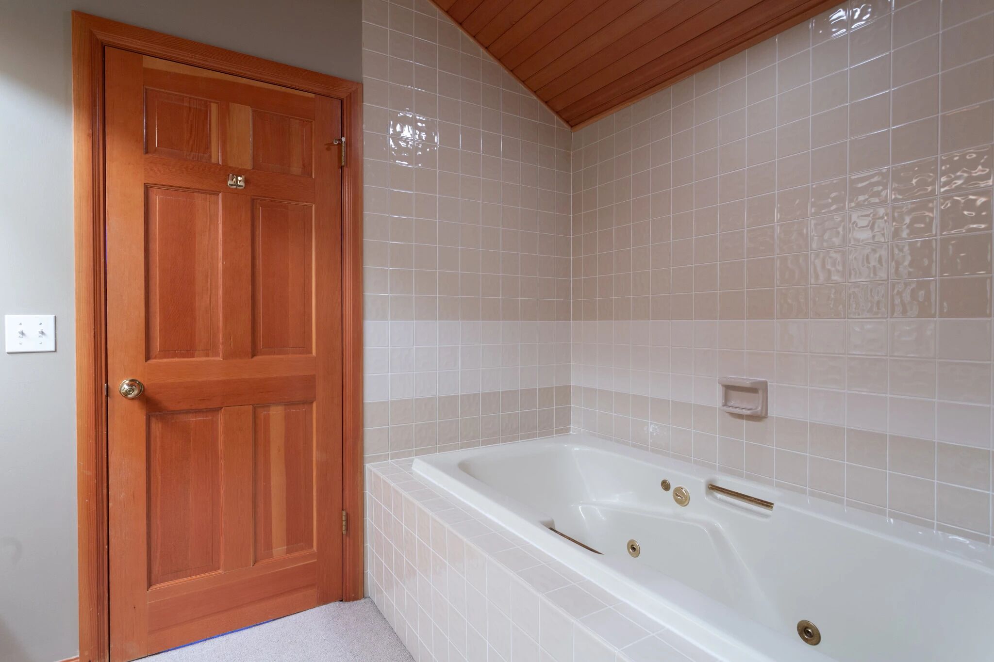 A bathroom with a wooden door, a white bathtub, and beige tiled walls.