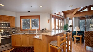 A modern kitchen with wooden cabinets, a granite countertop, and a built-in stove. There is a large window with a view of trees, and a cozy seating area with a green sofa and wooden chairs. - Ridge at Deer Valley Resort - Three Bedroom Condo with Hot Tub #16 3 Condo (Park City)