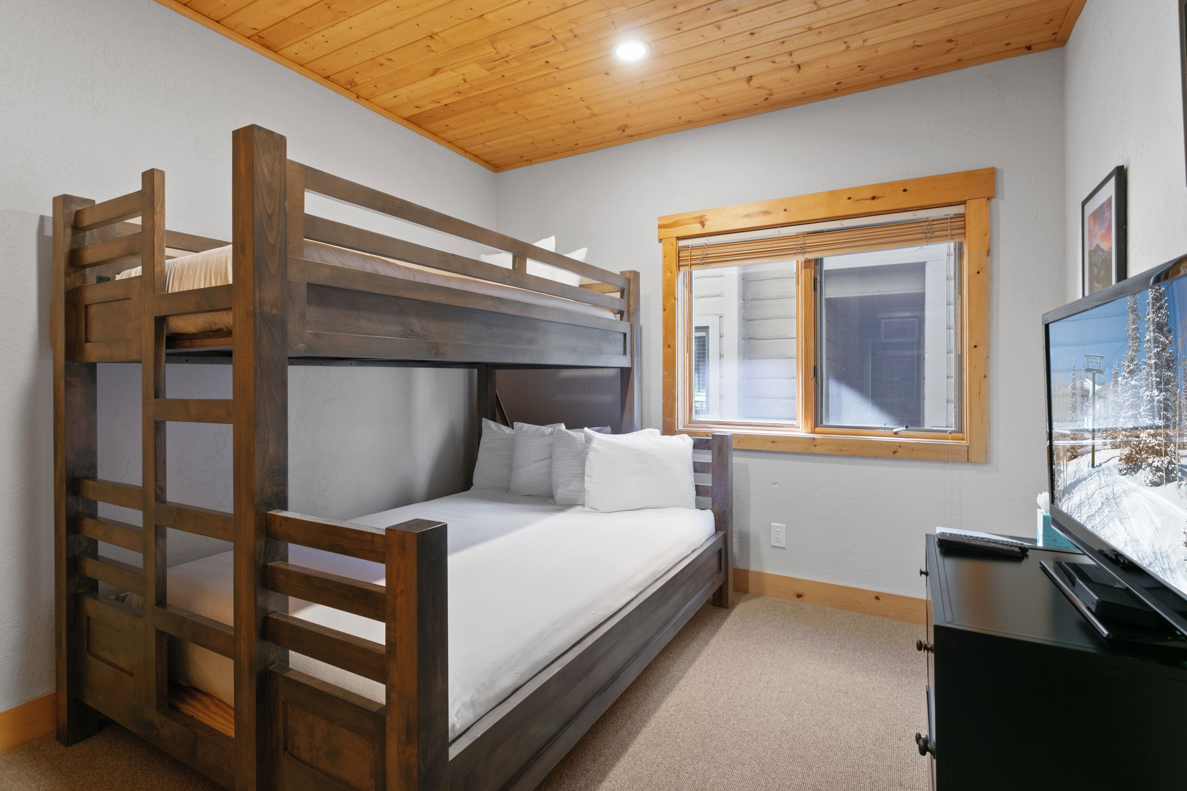 A bunk bed room with a wooden ceiling, a window with blinds, and a television on a dresser.