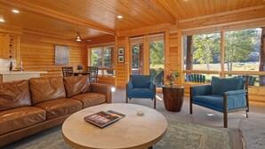 L'intérieur d'un chalet en bois est aménagé avec un canapé en cuir, deux fauteuils, une table basse ronde et de grandes fenêtres qui offrent une vue sur l'extérieur.
