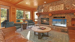 L’intérieur chaleureux d’un chalet en bois, avec une cheminée en pierre, des fauteuils confortables et une grande fenêtre donnant sur une forêt.