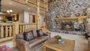 A cozy living room with a stone fireplace, wooden furniture, and a high wooden ceiling.